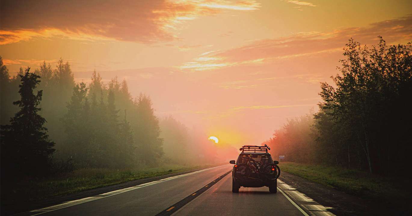 SUV-type vehicle driving into the orange-gold sunset on paved 2-lane road through the forest.