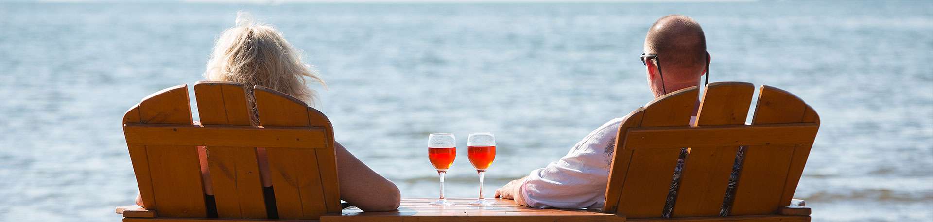 Man and woman sit in adjacent wooden chairs with 2 glasses of red wine on table and overlook Lake Mille Lacs.