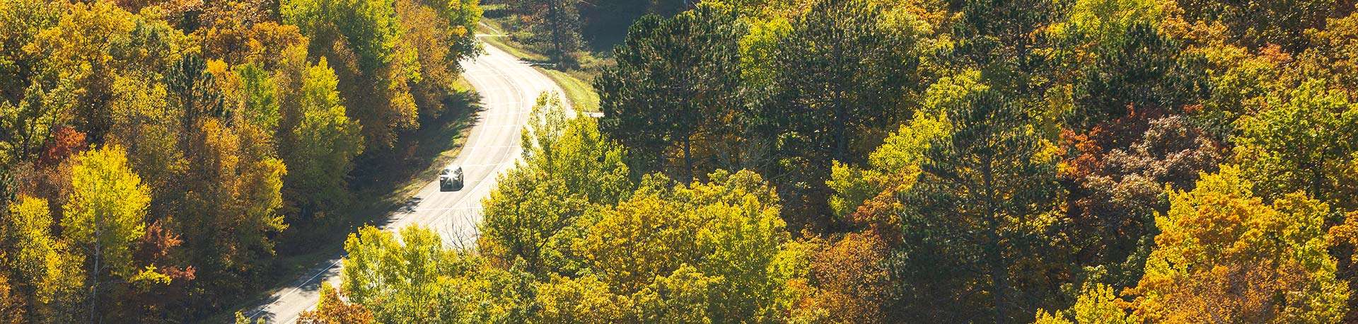 Aerial view of vehicle driving on 2-lane paved road through the autumn forest of orange, green, and gold.