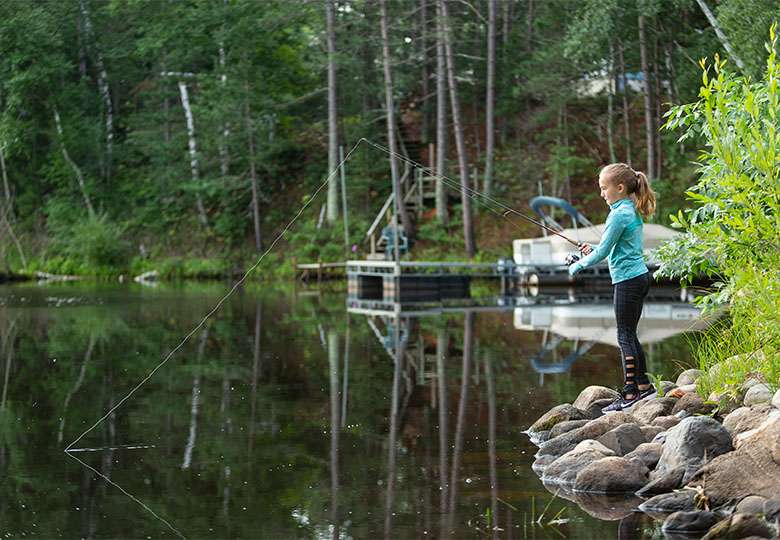 Girl in long-sleeved aqua shirt, black pants fishes from rocky shoreline, docked pontoon and cabin in background.