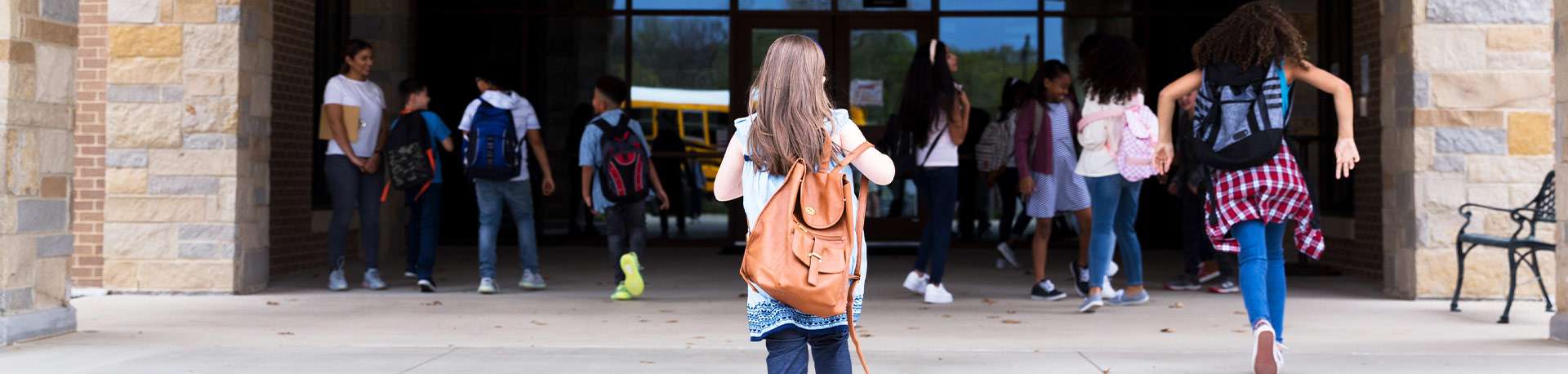 A dozen children approach the schoolhouse doors wearing backpacks.