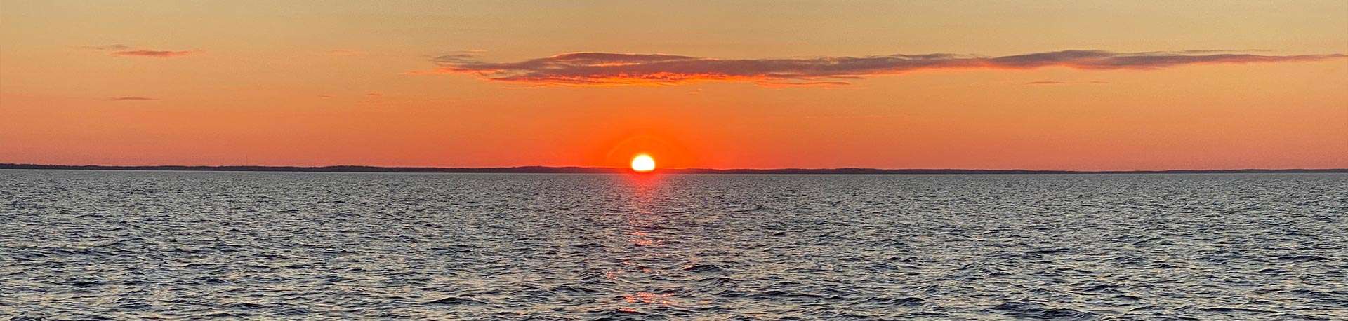 Orange sun setting over large lake, treeline in distance, one long wispy cloud high over the sun.